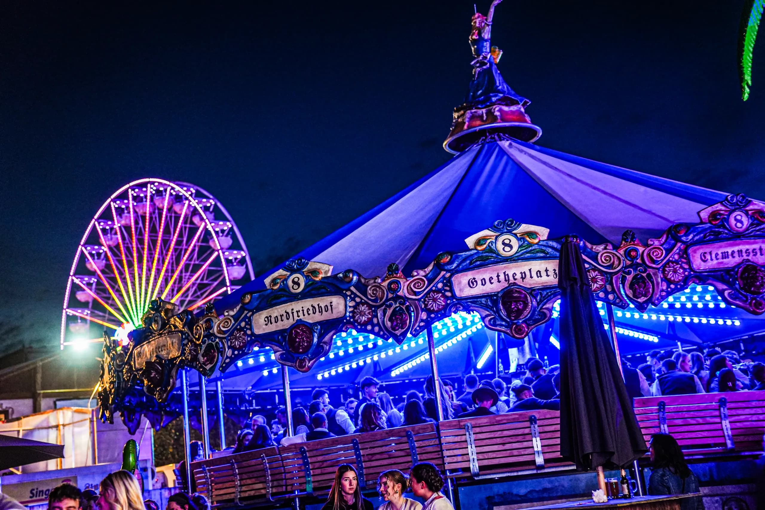 Das Karussell Linie 8 bei Nacht, blau und violett beleuchtet, daneben ein Riesenrad